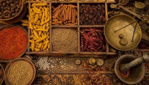 Top-down view of a bustling spice market stall, with overflowing wooden bins of turmeric, cinnamon sticks, star anise, cumin seeds, and dried chili peppers. Grains of rice scattered between the spices, tiny seed pods, and individual flakes of saffron. Weathered brass scales with weights, a mortar and pestle with residual colored powder. Intricate patterns on the wooden counter, cracks and grain visible. Warm, golden lighting, hyper-detailed texture of every spice, 8K resolution.