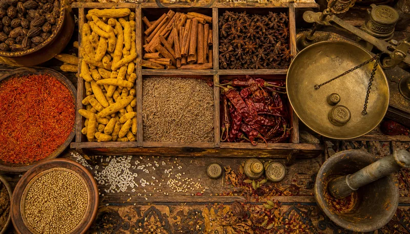 Top-down view of a bustling spice market stall, with overflowing wooden bins of turmeric, cinnamon sticks, star anise, cumin seeds, and dried chili peppers. Grains of rice scattered between the spices, tiny seed pods, and individual flakes of saffron. Weathered brass scales with weights, a mortar and pestle with residual colored powder. Intricate patterns on the wooden counter, cracks and grain visible. Warm, golden lighting, hyper-detailed texture of every spice, 8K resolution.