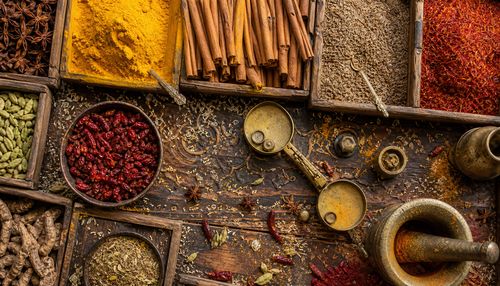 Top-down view of a bustling spice market stall, with overflowing wooden bins of turmeric, cinnamon sticks, star anise, cumin seeds, and dried chili peppers. Grains of rice scattered between the spices, tiny seed pods, and individual flakes of saffron. Weathered brass scales with weights, a mortar and pestle with residual colored powder. Intricate patterns on the wooden counter, cracks and grain visible. Warm, golden lighting, hyper-detailed texture of every spice, 8K resolution.